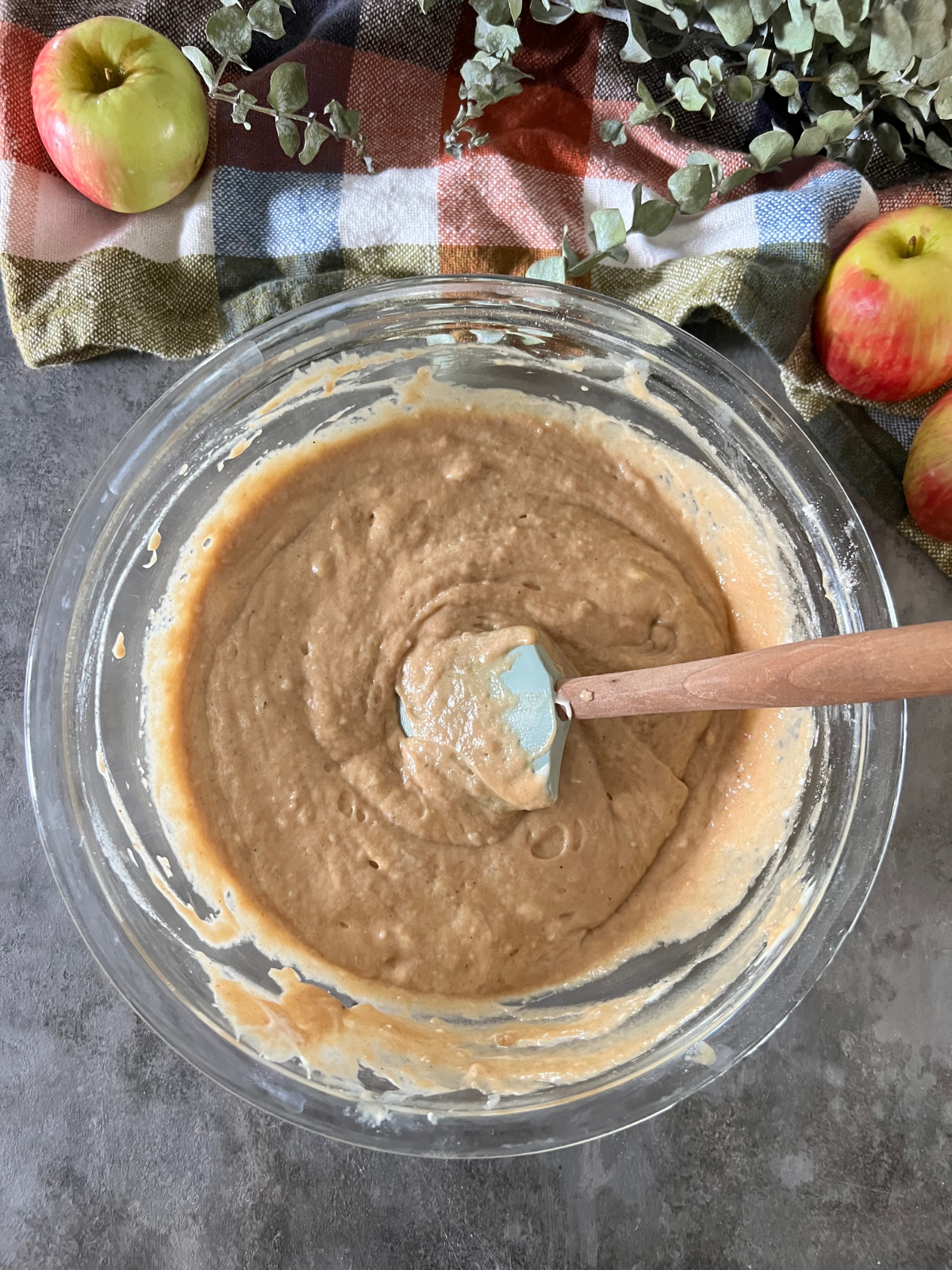 A glass mixing bowl sits with a rubber spatula with all the apple cider donut batter in it.