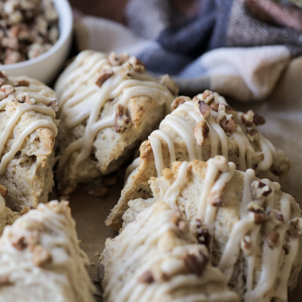 Several maple pecan sourdough scones sit on a cutting board with maple glaze and chopped pecans sprinkled over the top of them. A kitchen towel and bowl of chopped pecans sit in the background.