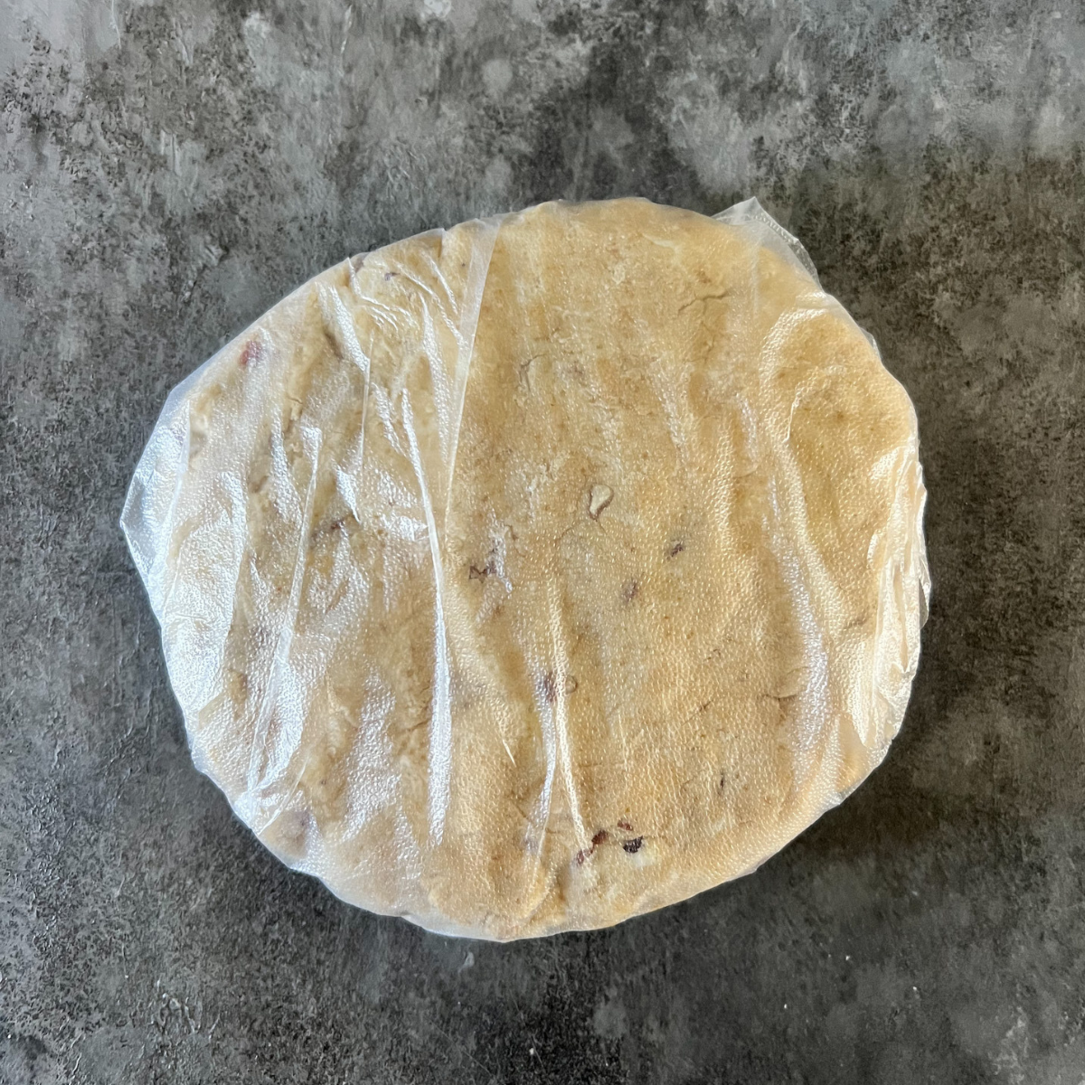 The disc of sourdough maple pecan scone dough sits on the counter and is wrapped in plastic wrap.
