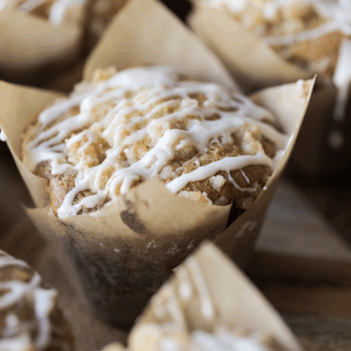 A closeup of a sourdough vanilla bean muffin with crumble topping and vanilla bean icing. It is surrounded by other muffins.