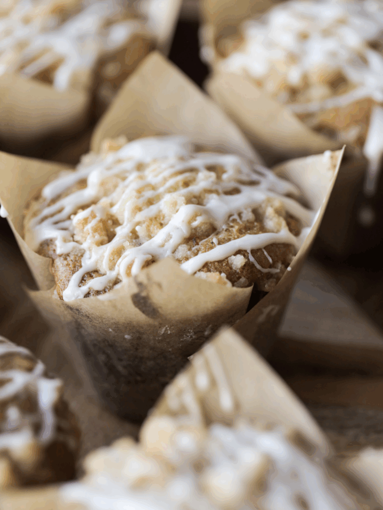 A closeup of a sourdough vanilla bean muffin with crumble topping and vanilla bean icing. It is surrounded by other muffins.