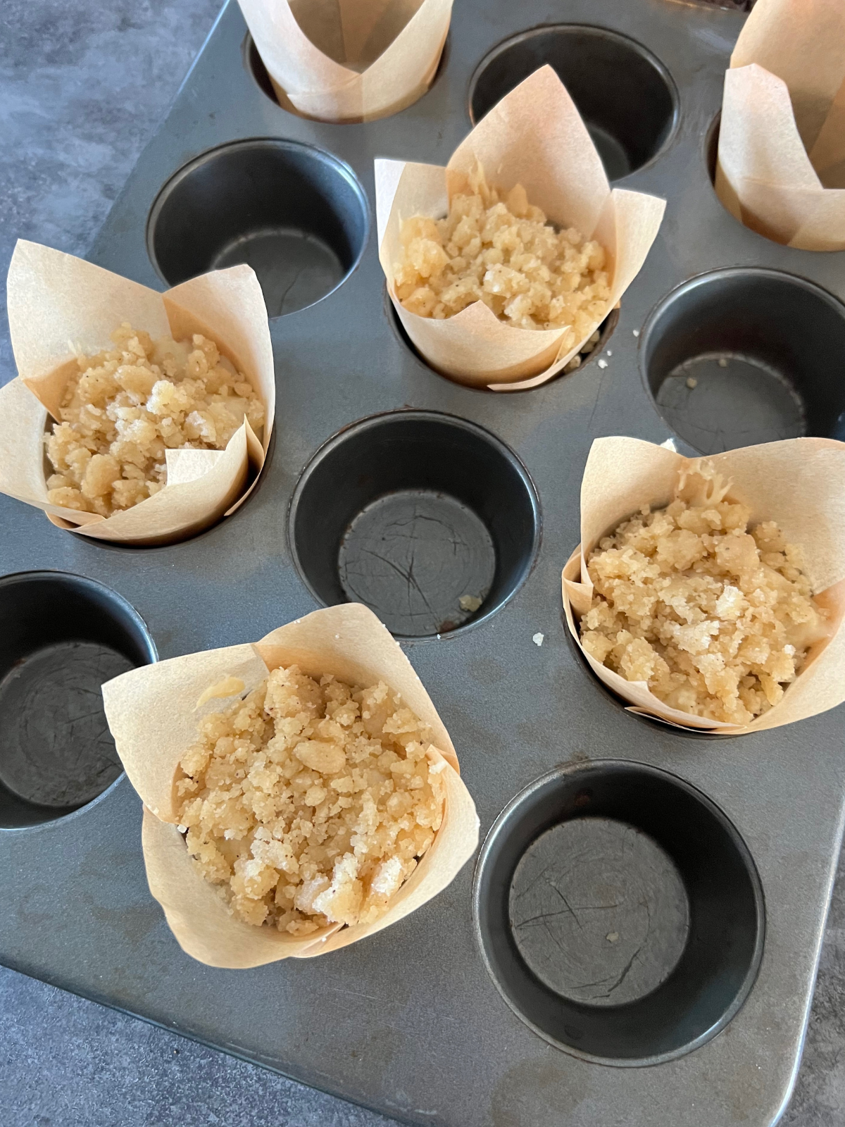 A muffin tin holds the cupcake batter and crumble. The pan has every other hole filled and the cupcakes have liners. 