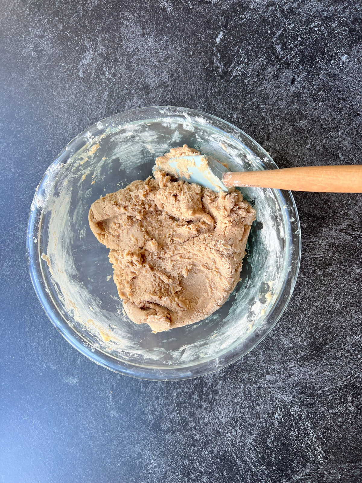 A glass mixing bowl with a rubber spatula sit on a counter with the sourdough snickerdoodle cookie dough in it.