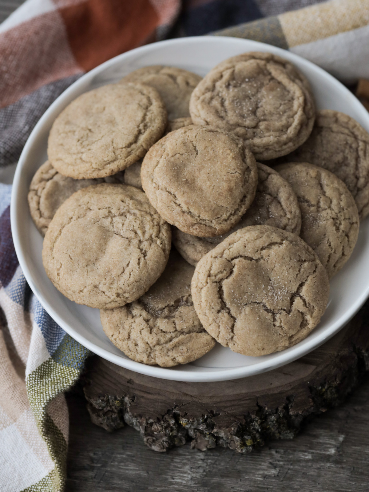 A plate of incredibly chewy and soft sourdough discard snickerdoodles sit on a wooden slab with a plaid kitchen towel in the background.