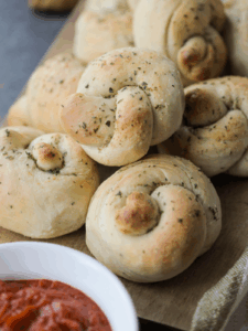 A closeup of sourdough garlic and herb knots with a bowl of marinara in the foreground.