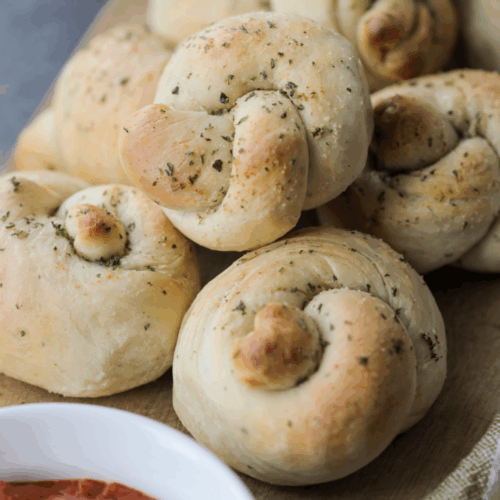 A closeup of sourdough garlic and herb knots with a bowl of marinara in the foreground.