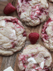 Several sourdough discard raspberry white chocolate cookies surrounded by raspberries and chopped white chocolate.