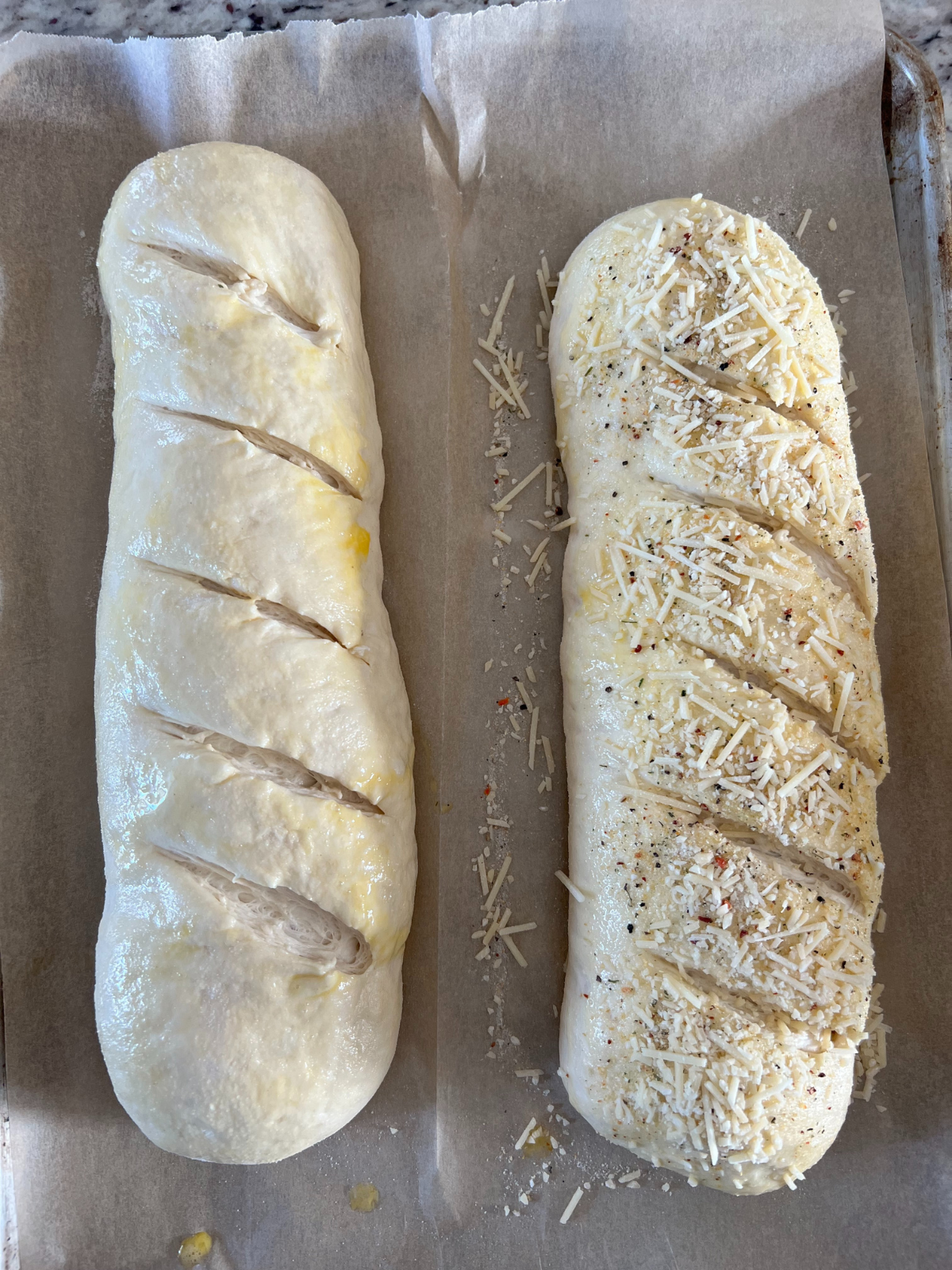The risen sourdough French bread is shown on the baking sheet after being scored diagonally. Both loaves have been brushed with egg wash. One loaf has Italian seasoning and shredded parmesan cheese. 