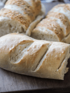 Three half loaves of sourdough french bread sit on a wooden cutting board. Two of them are italian herbs and cheese bread.