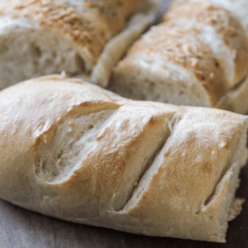 Three half loaves of sourdough french bread sit on a wooden cutting board. Two of them are italian herbs and cheese bread.