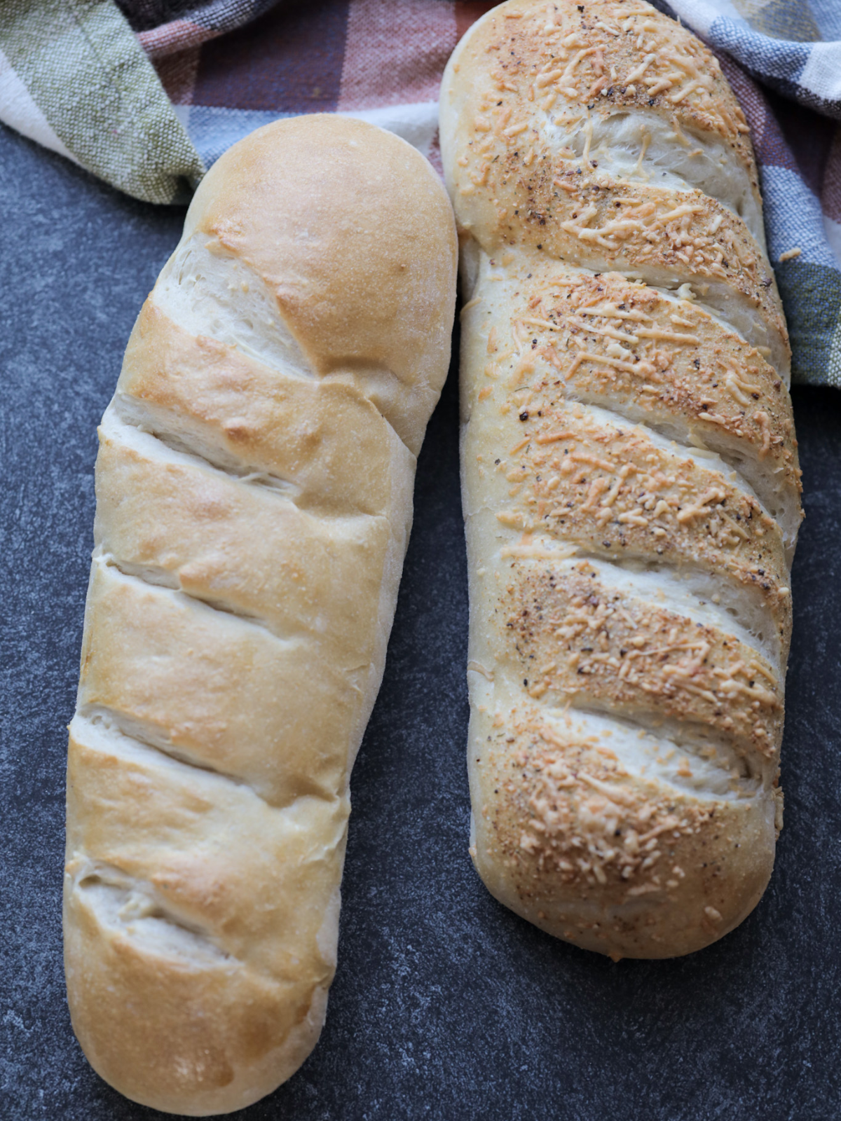 Two loaves of sourdough French bread sit. One loaf has Italian herbs and shredded parmesan cheese. 