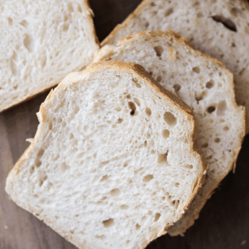 Three slices of soft sourdough sandwich bread sit next to the loaf of bread on a wooden cutting board.