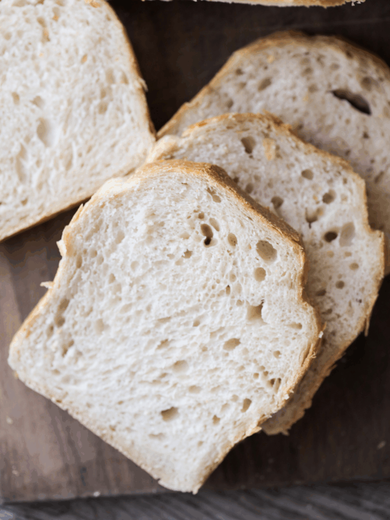 Three slices of soft sourdough sandwich bread sit next to the loaf of bread on a wooden cutting board.