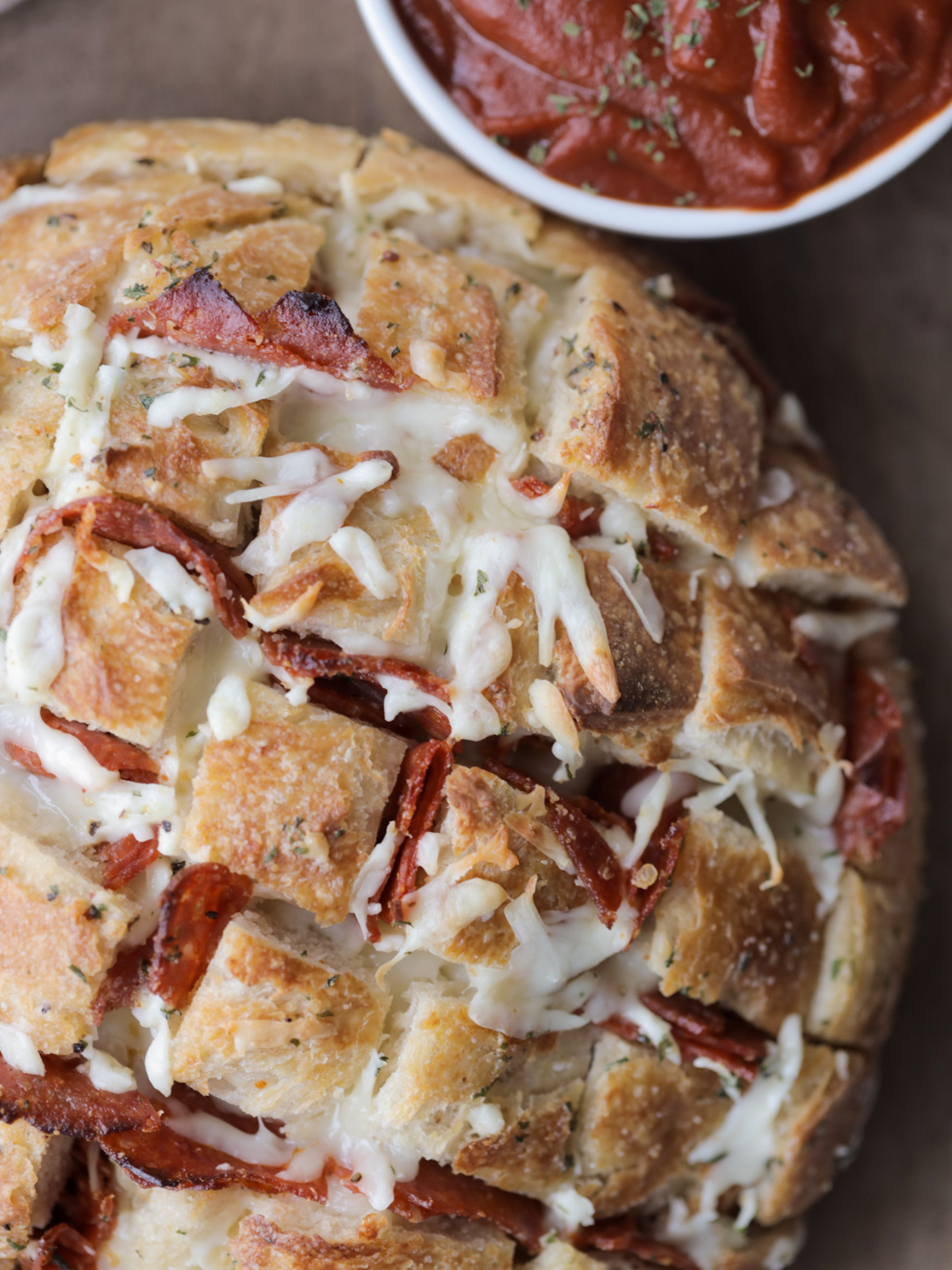 The freshly baked cheesy mozzarella and pepperoni sourdough loaf sits on a wooden cutting board aside a bowl of marinara sauce. 