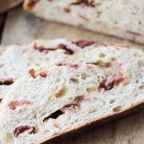 Two slices of sourdough white chocolate strawberry bread sit on a wooden cutting board with half of the loaf in the background.