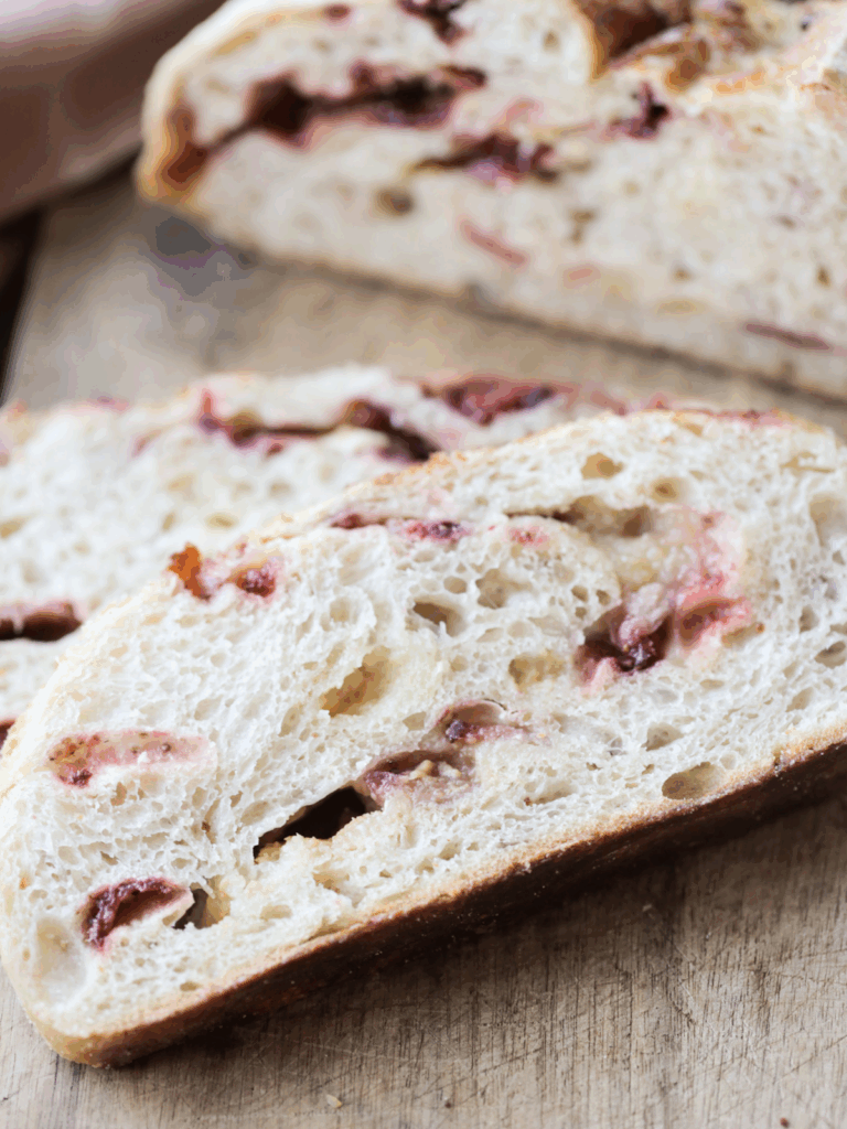 Two slices of sourdough white chocolate strawberry bread sit on a wooden cutting board with half of the loaf in the background.