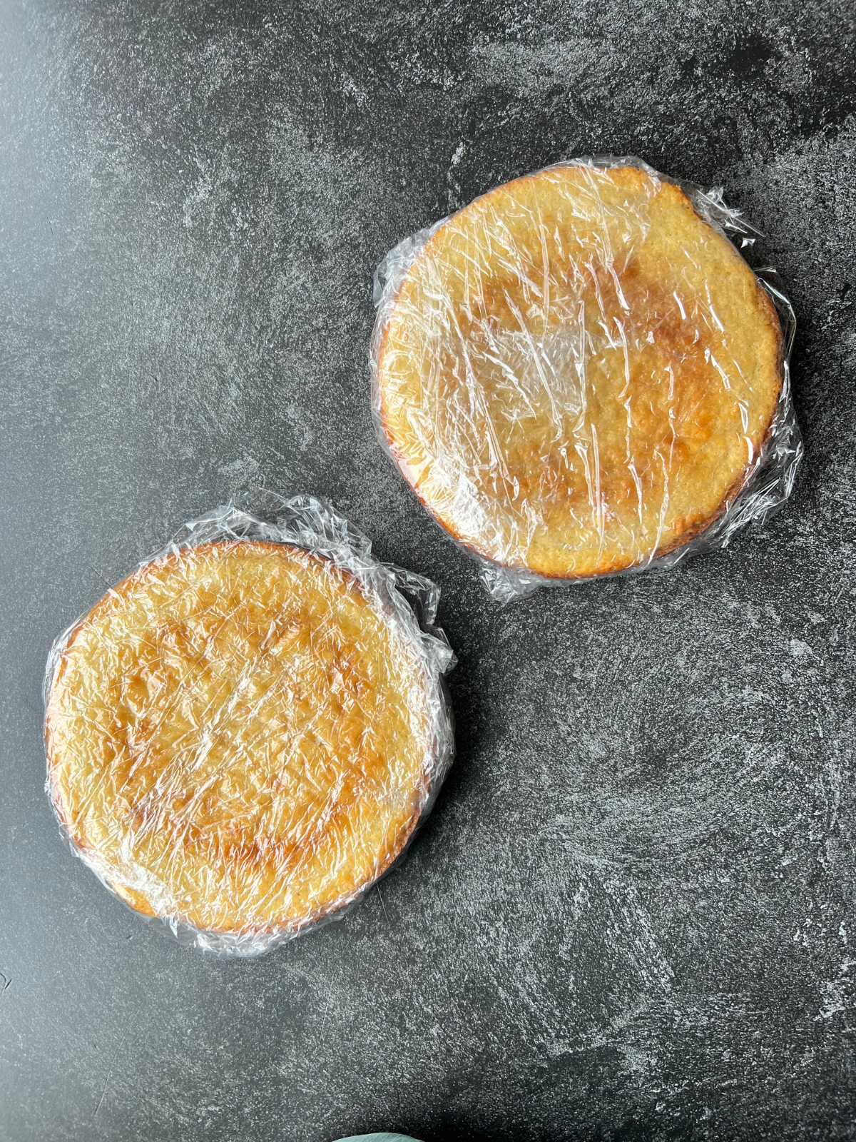 Two baked and cooled sourdough vanilla cakes sit wrapped in plastic wrap. This preserves their moisture if you bake the day ahead of time.