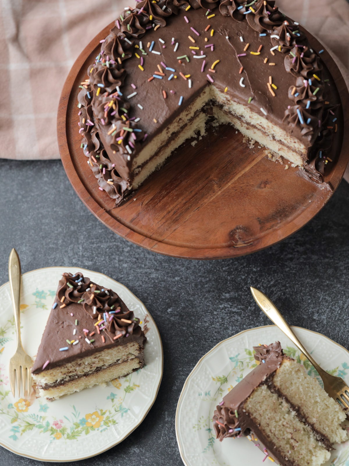 The sourdough vanilla birthday cake sits on a wooden cake stand. Two slices have been taken out and each sits on a floral plate with forks.