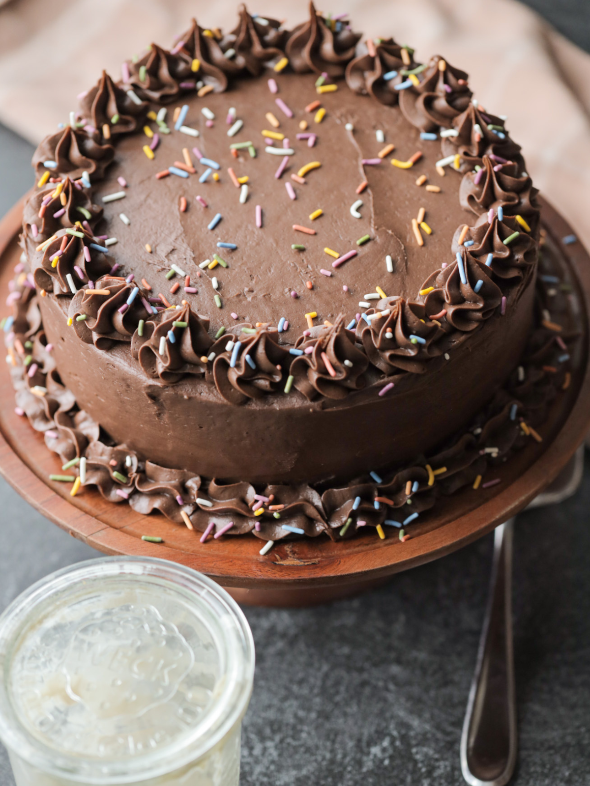 The full sourdough vanilla birthday cake with chocolate cream cheese frosting and sprinkles sits on a wooden cake stand. A jar of sourdough discard sits in front of it.