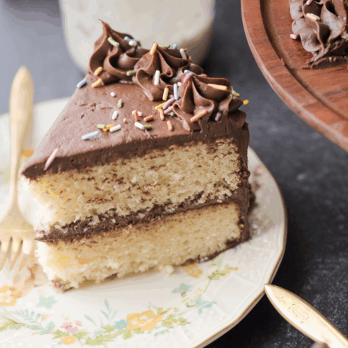 One slice of a two-layered sourdough vanilla birthday cake with chocolate frosting and sprinkles sits on a plate with a fork.
