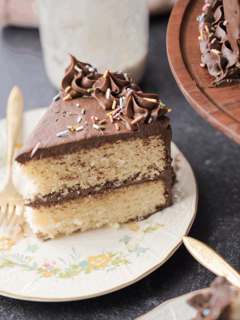 One slice of a two-layered sourdough vanilla birthday cake with chocolate frosting and sprinkles sits on a plate with a fork.