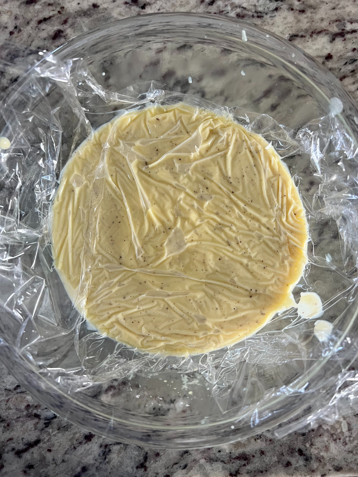 The vanilla bean custard sits in a glass mixing bowl with plastic wrap over it. 