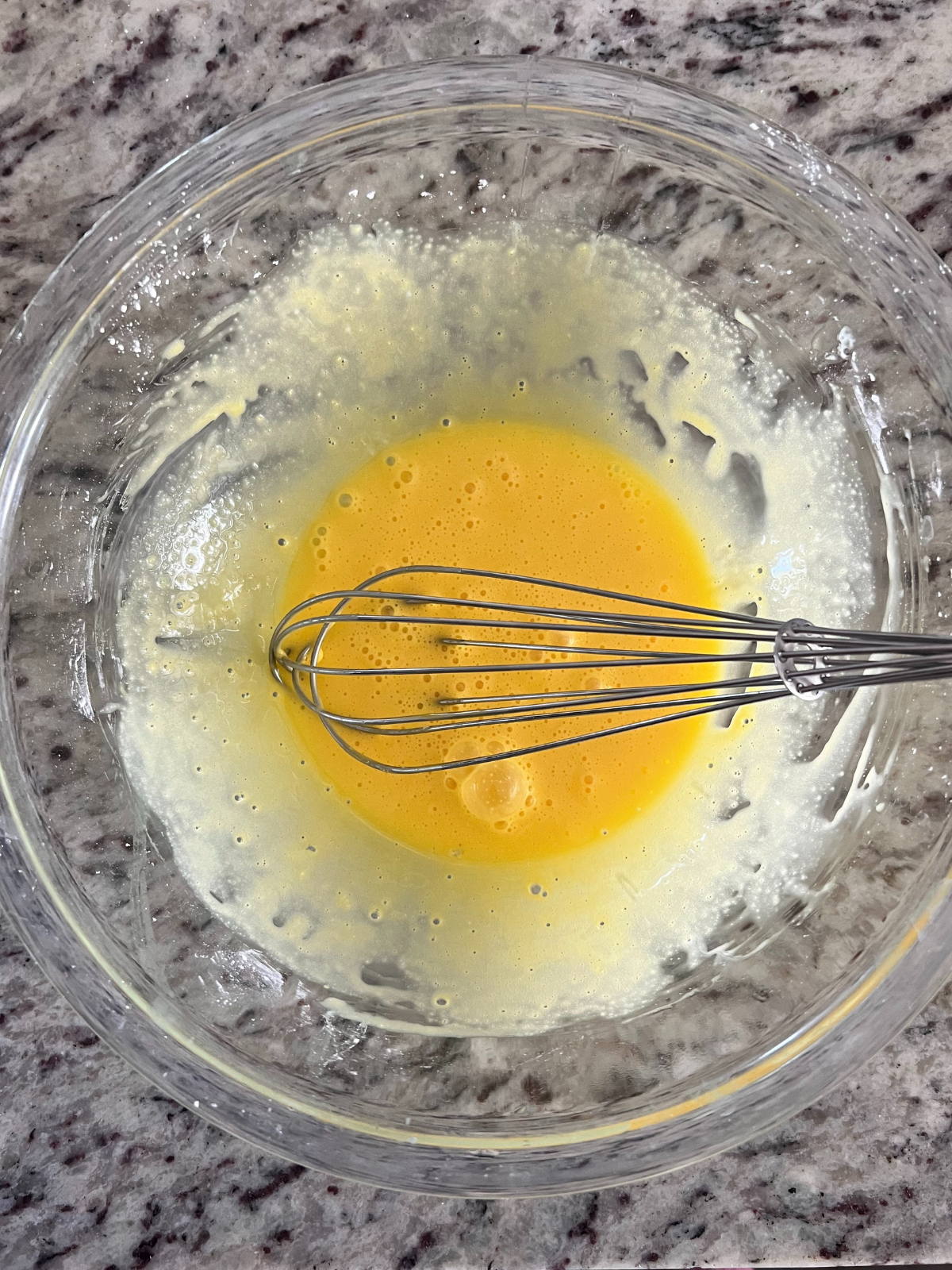 A glass mixing bowl holds whisked together egg yolks and cornstarch. 