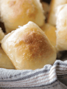 One sourdough brioche dinner roll shown closeup with several in the background. They sit in a basket lined with a kitchen towel.