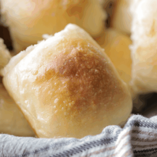 One sourdough brioche dinner roll shown closeup with several in the background. They sit in a basket lined with a kitchen towel.