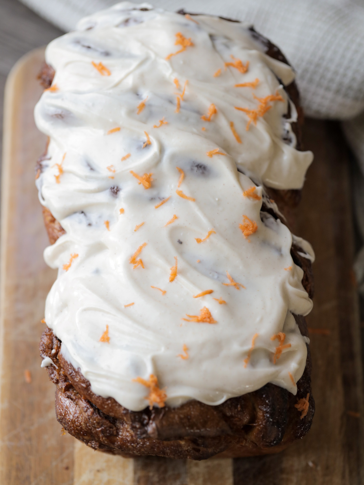 Sourdough carrot cake babka sits on a wooden cutting board with cinnamon cream cheese frosting and grated carrots over the top.