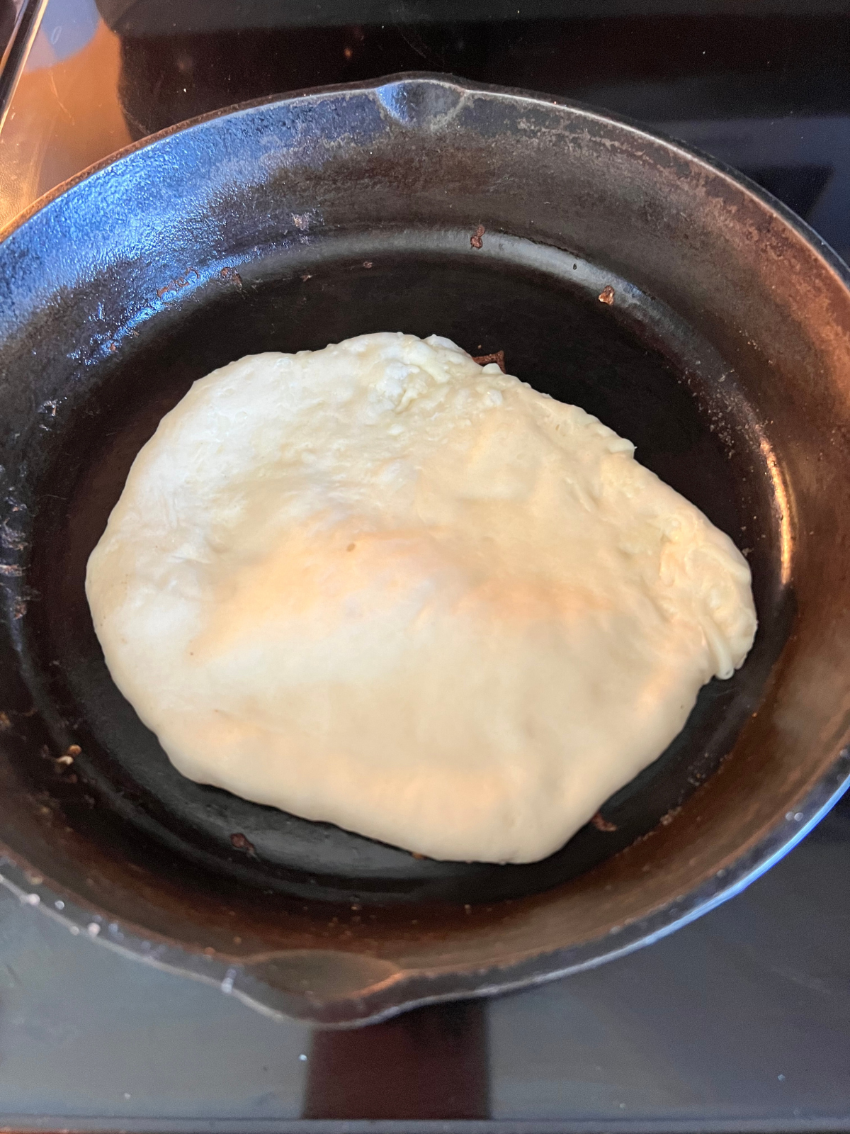 The rolled out sourdough naan dough is cooking on the cast-iron skillet. It is bubbling up which shows it is ready to be flipped and cooked on the other side.
