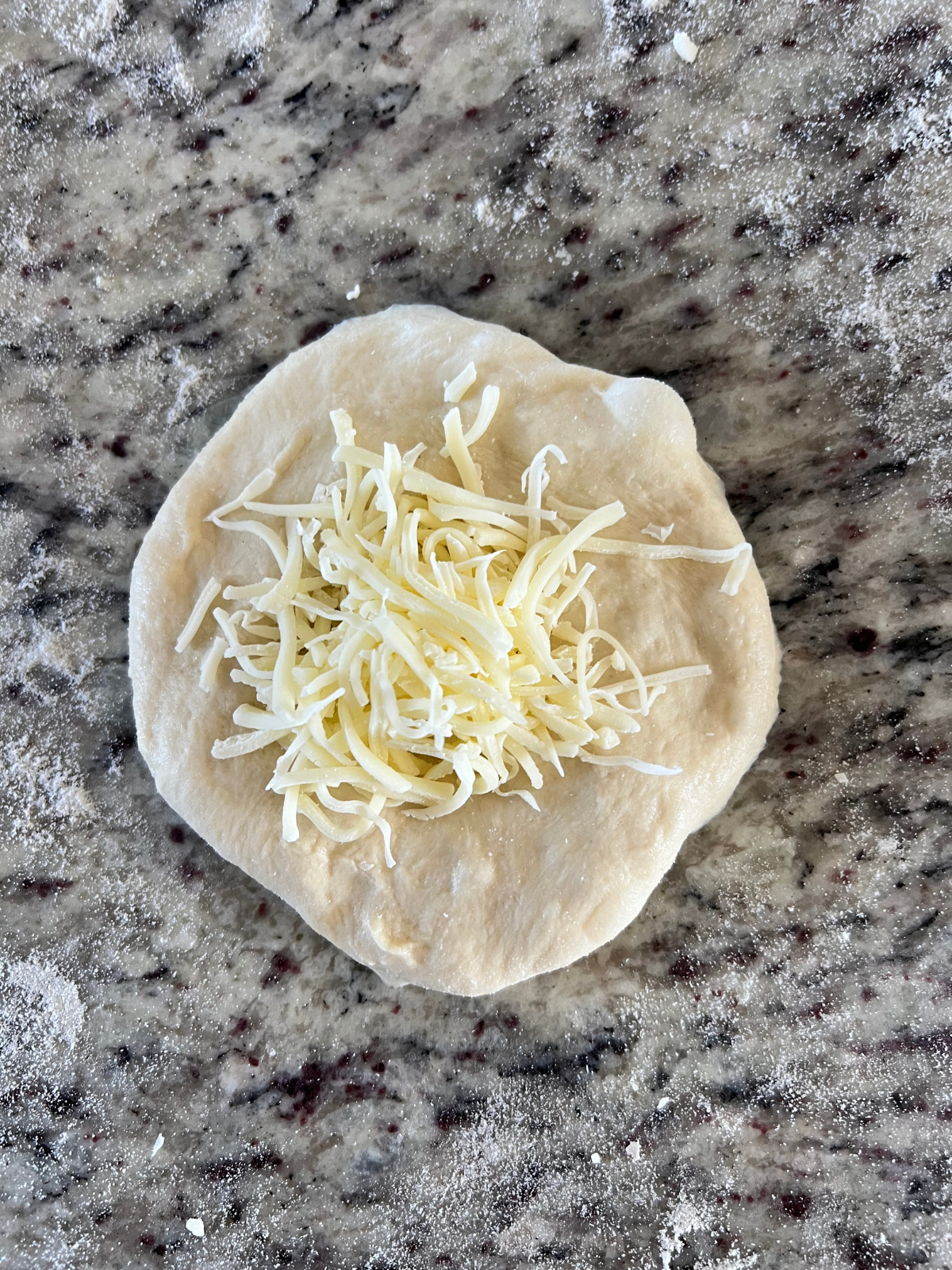 A flattened sourdough naan dough ball is shown with shredded mozzarella cheese on top.