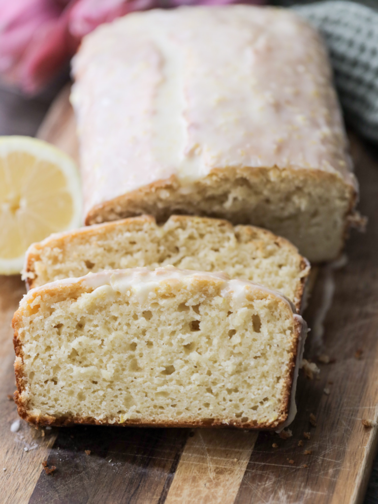 Sourdough lemon pound cake sits on a wooden cutting board behind two slices of the cake.
