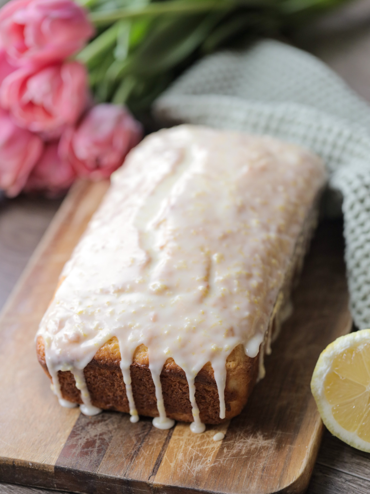 A loaf of sourdough discard lemon pound cake sits on a wooden cutting board. Lemon glaze is drizzled over it.