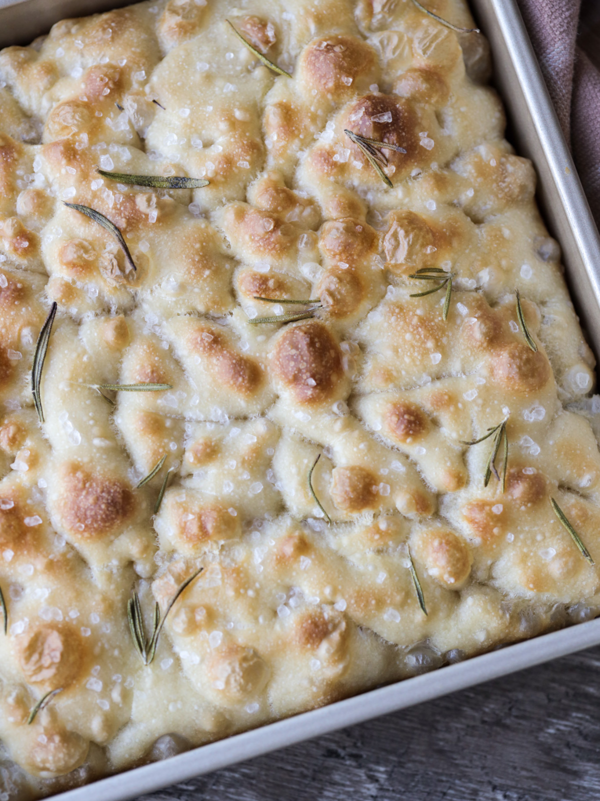 A closeup of the sourdough focaccia bread with flaky sea salt and rosemary sprigs is shown here.