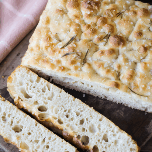 Sourdough focaccia sits on a wooden cutting board. Two slices have been cut out, showing the airy and bubbly interior of the bread.