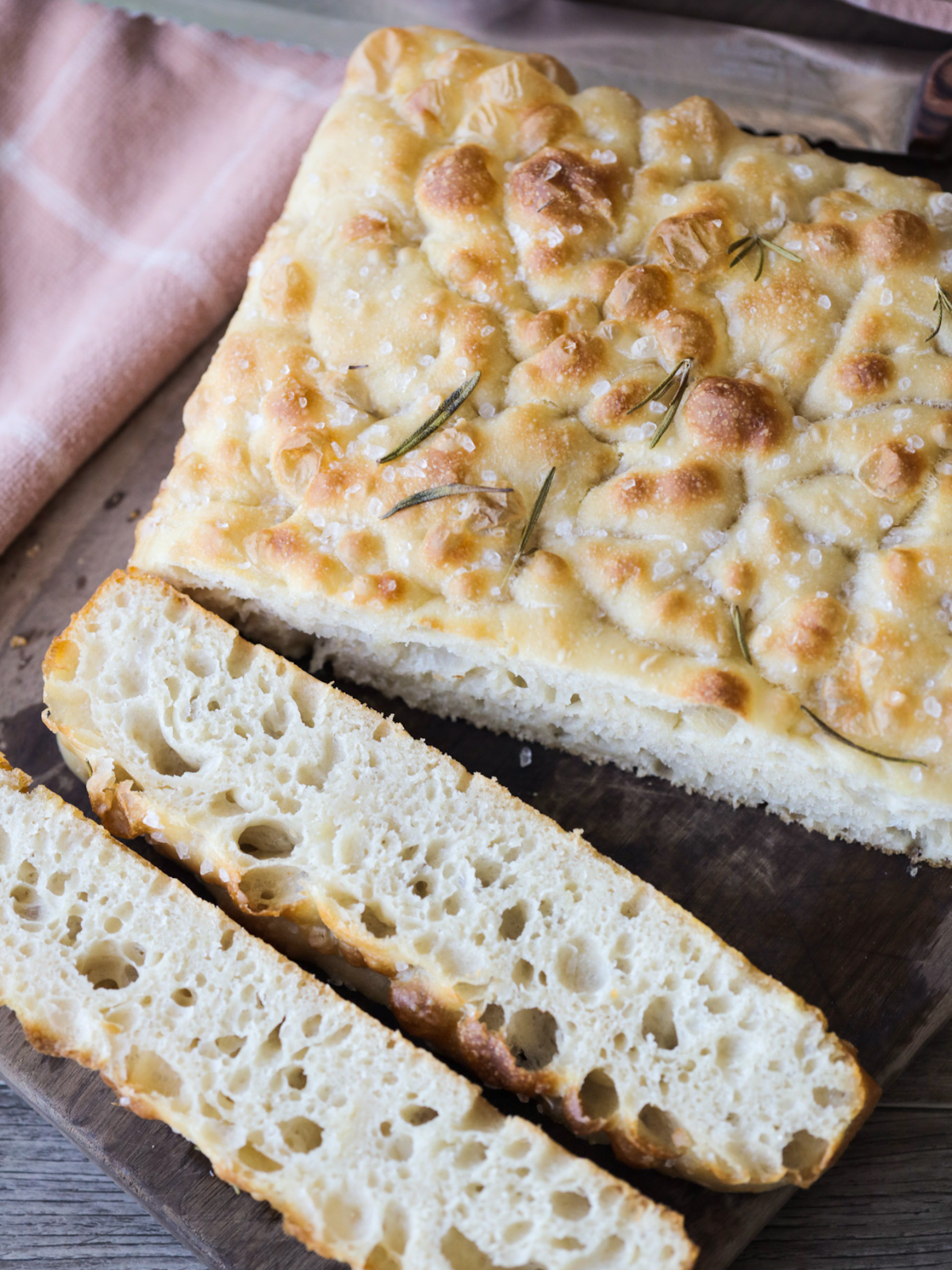 Sourdough focaccia sits on a wooden cutting board. Two slices have been cut out, showing the airy and bubbly interior of the bread.