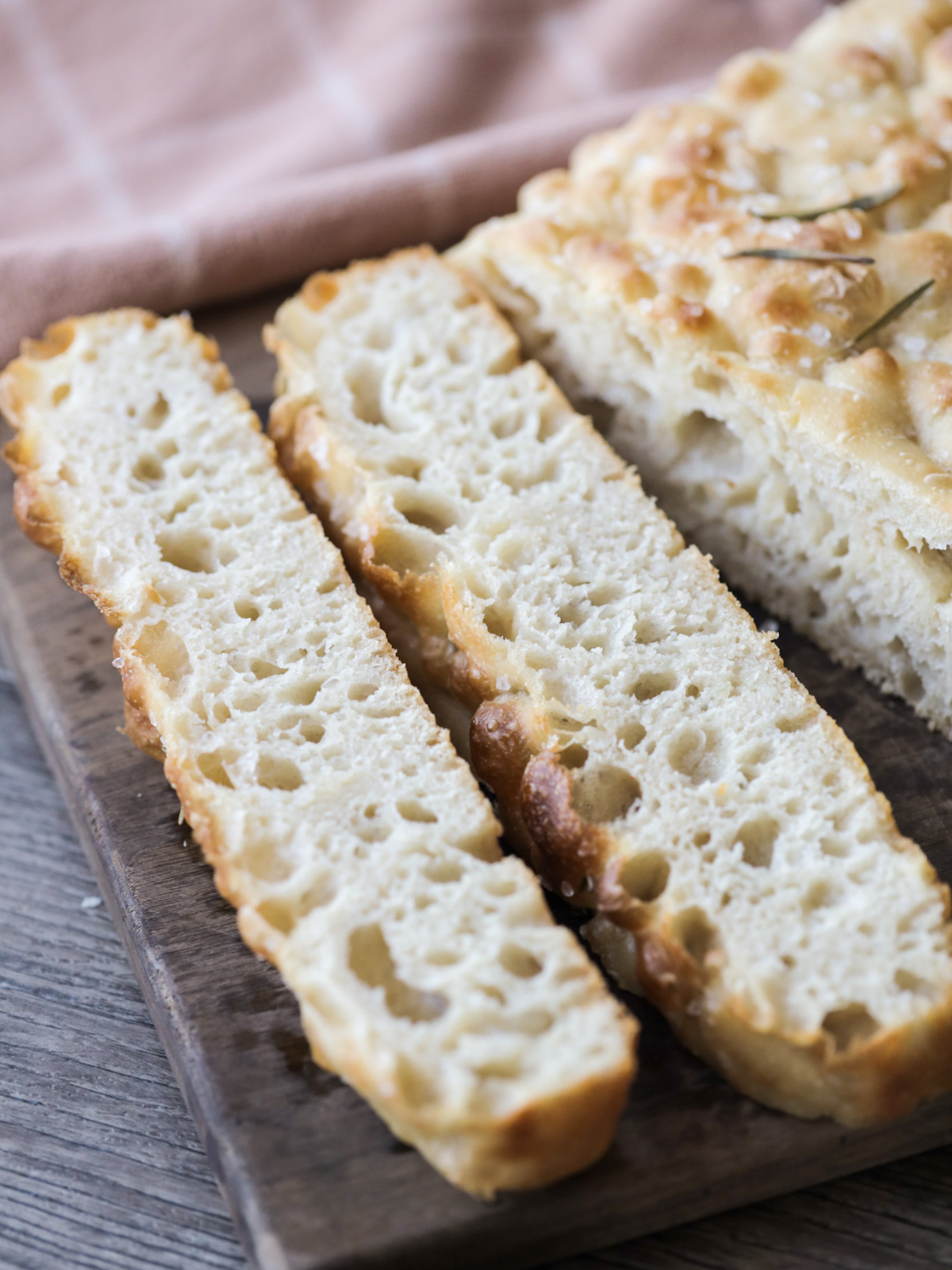 A closeup of the bubbly and airy sourdough focaccia slices are shown on a wooden cutting board. 