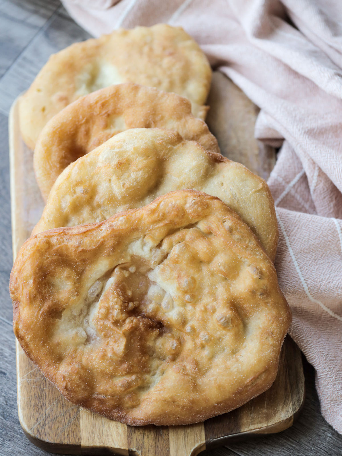 Several sourdough fry bread sit on a wooden cutting board with a kitchen towel to the side.