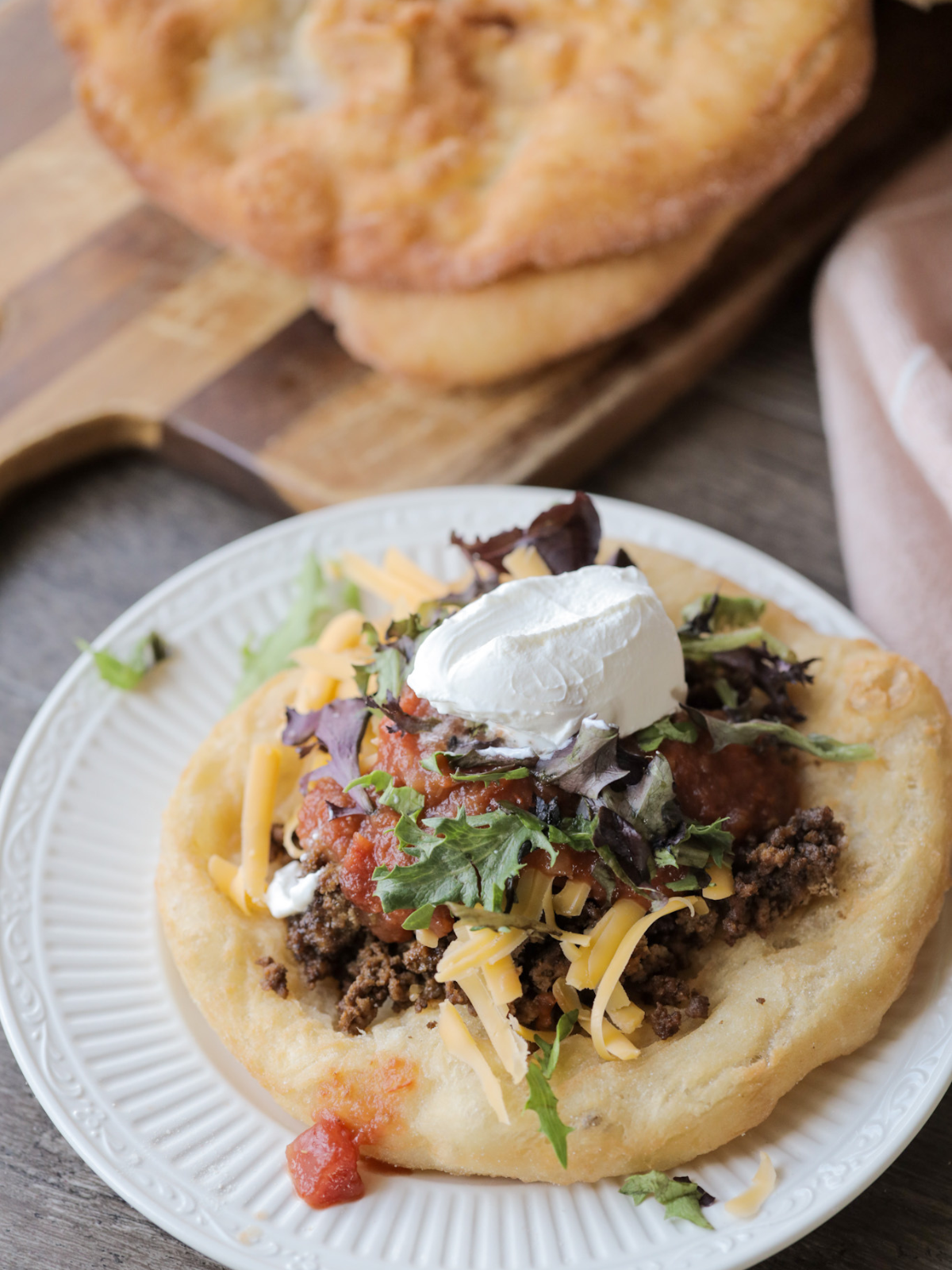 A delicious sourdough fry bread taco with all the fixings sit on a plate with a stack of sourdough fry bread sitting in the background.