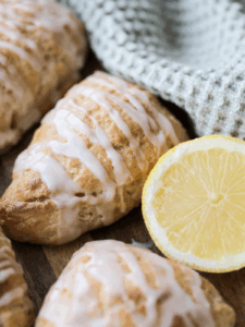 A closeup of a lemon sourdough scone with lemon glaze is shown next to a lemon and several other scones in the background.