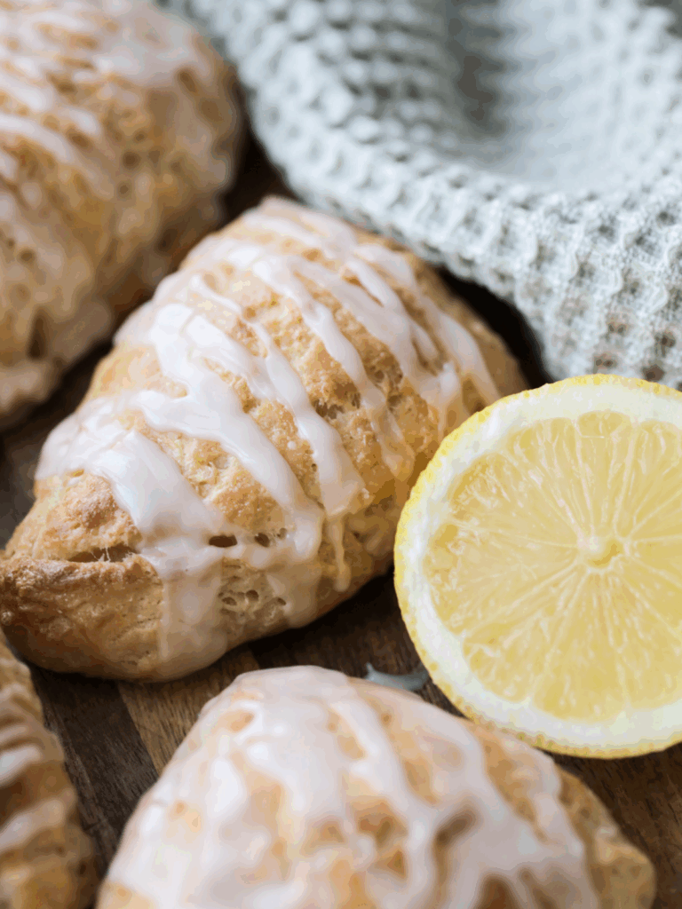 A closeup of a lemon sourdough scone with lemon glaze is shown next to a lemon and several other scones in the background.