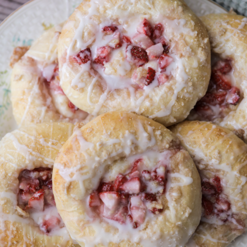 Several sourdough discard strawberry cream cheese buns sitting on a floral plate. They are covered in streusel and icing.