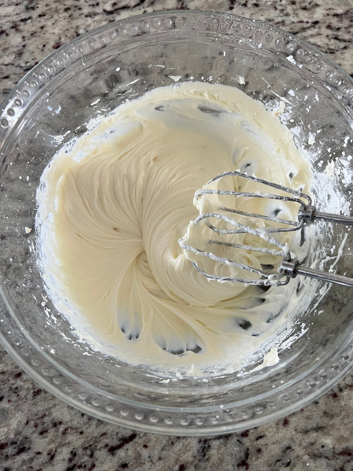 The cream cheese filling sits in a glass mixing bowl with hand mixer beaters sitting to the side of the bowl.