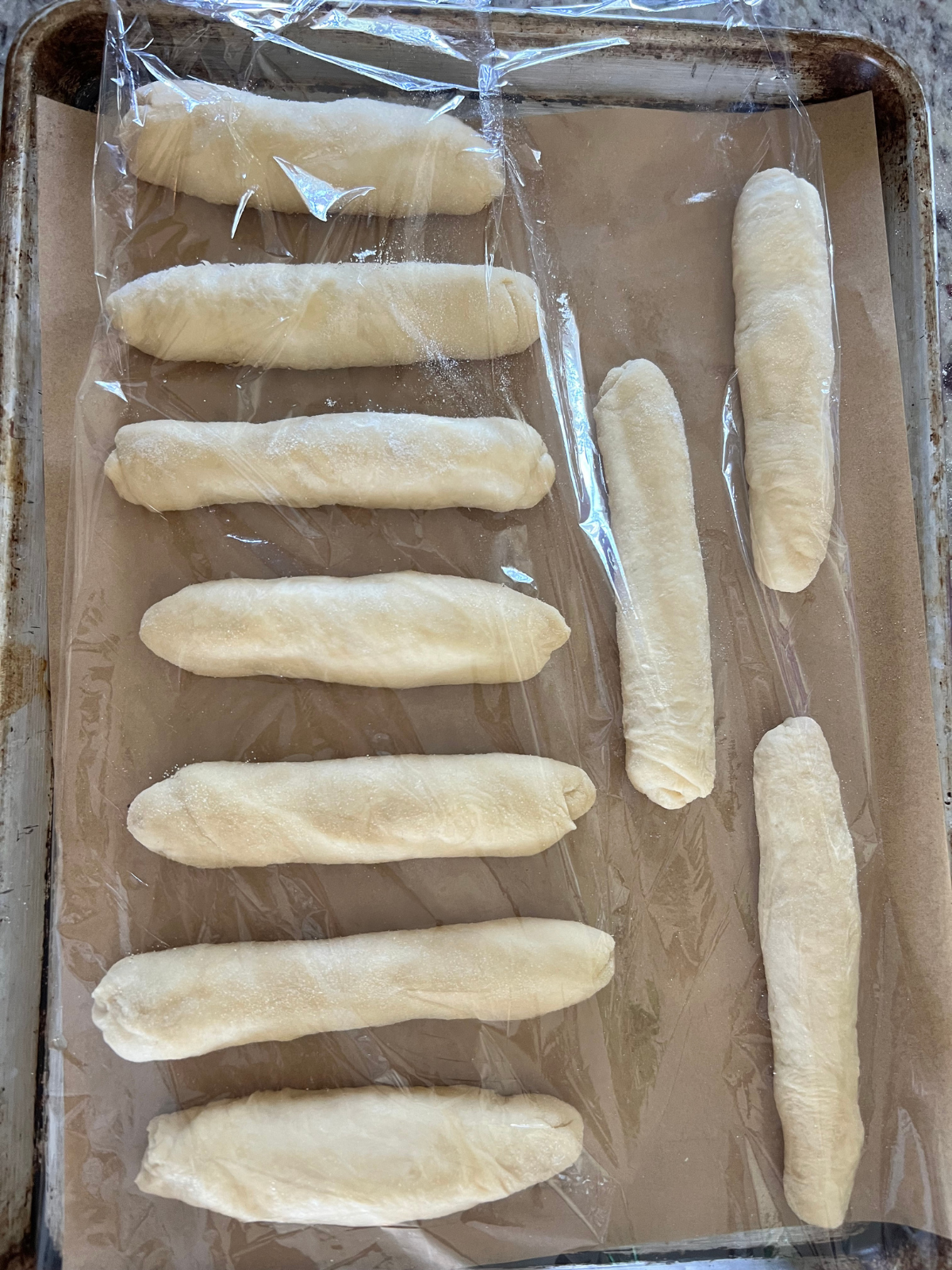 All ten sourdough hot dog buns are on a parchment paper-lined baking sheet with greased plastic wrap covering them. They will have their second rise for about 1-4 hours.