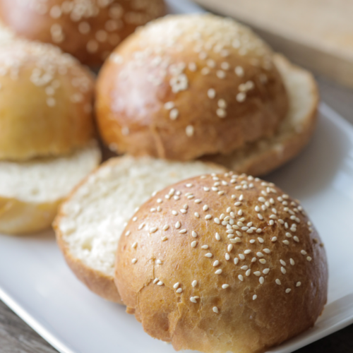 A closeup of one sourdough brioche burger bun that was cut in half. It is topped with sesame seeds. Several more burger buns rest in the background.