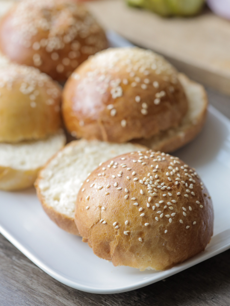 A closeup of one sourdough brioche burger bun that was cut in half. It is topped with sesame seeds. Several more burger buns rest in the background.