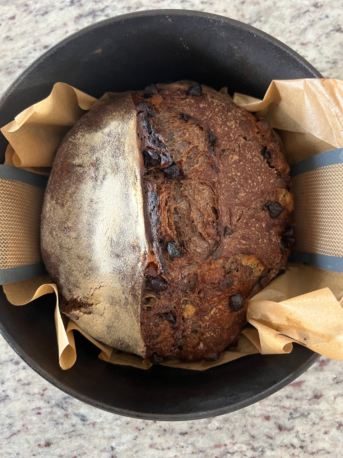 The sourdough chocolate and coffee loaf sits in a Dutch oven fresh out of the oven. It rests on both parchment paper and a bread sling.