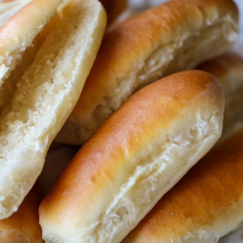 Several soft sourdough hot dog buns rest in a basket. One bun is cut open to show the soft, fluffy interior.