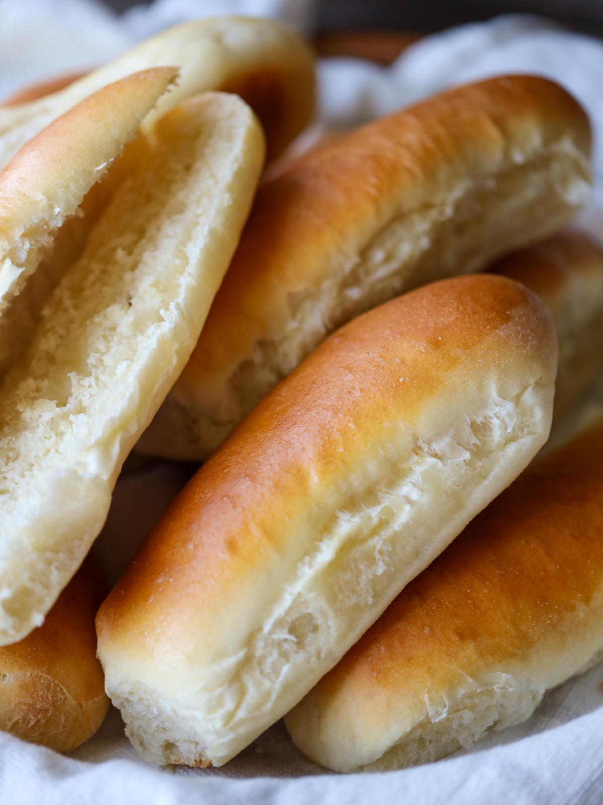 Several soft sourdough hot dog buns rest in a basket. One bun is cut open to show the soft, fluffy interior.